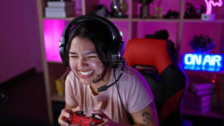 A young hispanic woman enjoys gaming at night in an indoor room illuminated with purple lighting, wearing headphones and smiling.