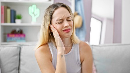 Téléchargez les photos : Jeune femme blonde assise sur un canapé souffrant de mal de dents à la maison - en image libre de droit