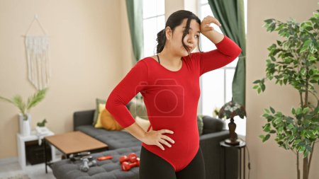 Foto de Mujer joven hispana en ejercicio rojo en el interior, descansando con una mano en la cadera y limpiando el sudor de su frente - Imagen libre de derechos