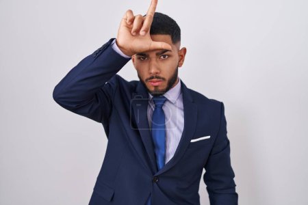 Photo for Young hispanic man wearing business suit and tie making fun of people with fingers on forehead doing loser gesture mocking and insulting. - Royalty Free Image