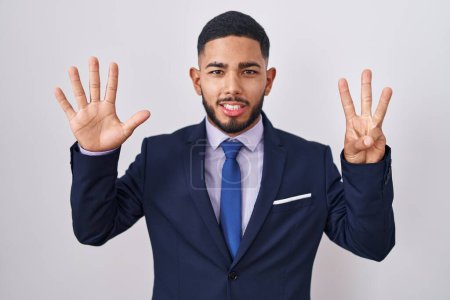 Foto de Joven hombre hispano usando traje de negocios y corbata mostrando y señalando con los dedos número ocho mientras sonríe confiado y feliz. - Imagen libre de derechos