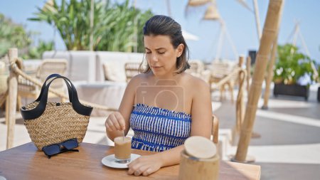 Foto de Mujer hispana joven revolviendo café sentado en la mesa en la terraza soleada del restaurante - Imagen libre de derechos