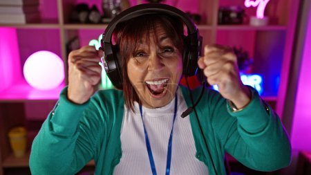 Foto de Una alegre mujer hispana de mediana edad disfruta jugando por la noche usando auriculares en una colorida habitación interior. - Imagen libre de derechos