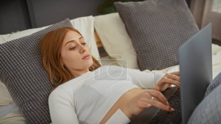 A young caucasian redhead woman lounges in bed using a laptop in a cozy, modern bedroom setting.