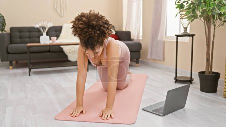 Photo for A young woman in a pink bodysuit practices yoga in a well-lit living room with a laptop on the floor. - Royalty Free Image