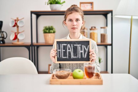 Photo for Relaxed yet serious, young blonde woman catches a healthy breakfast at home, with simple natural expression. looking straight at the camera, holding a blackboard. - Royalty Free Image
