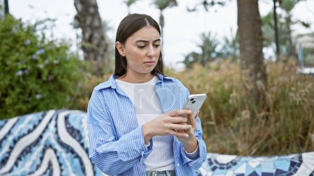 Photo for A concerned young hispanic woman browses her smartphone in a lush park. - Royalty Free Image