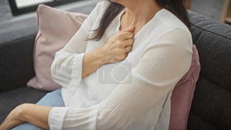 Photo for Middle-aged woman clutching chest in discomfort while seated on a sofa indoors, conveying potential health issues. - Royalty Free Image