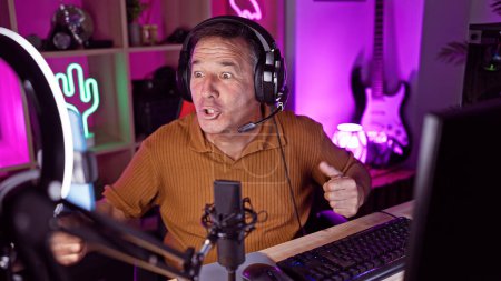 Surprised man wearing headphones in a colorful gaming room with microphone and computer.