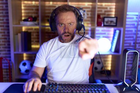 Foto de Hombre de mediana edad con barba jugando videojuegos con auriculares apuntando disgustado y frustrado a la cámara, enojado y furioso con usted - Imagen libre de derechos