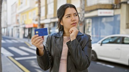 Pensive young hispanic woman holding credit card on city street, embodying urban lifestyle and financial contemplation.