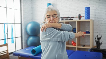 Senior woman stretching in a physiotherapy rehab clinic room with exercise equipment indoors.