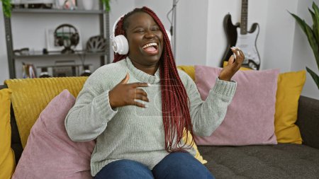 Foto de Alegre mujer negra con trenzas con auriculares finge tocar la guitarra en casa en un sofá acogedor - Imagen libre de derechos