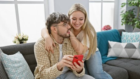 Ein Liebespaar genießt die gemeinsame Zeit im Haus, wobei die Frau dem faszinierten Mann in einem gemütlichen Wohnzimmer etwas auf dem Smartphone zeigt.