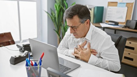 Téléchargez les photos : Un homme d'âge moyen dans des lunettes éprouvant de l'inconfort à son bureau, serrant sa poitrine dans une détresse potentielle pour la santé - en image libre de droit
