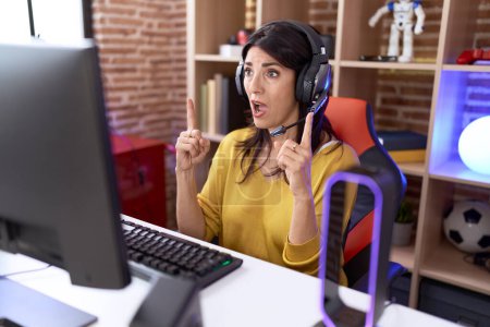 Foto de Mujer hispana de mediana edad jugando videojuegos usando auriculares asombrada y sorprendida mirando hacia arriba y señalando con los dedos y los brazos levantados. - Imagen libre de derechos