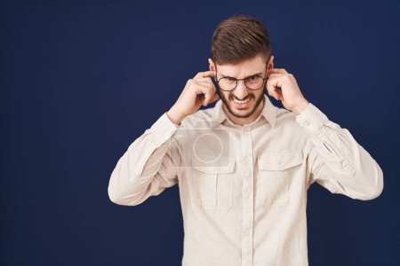 Photo for Hispanic man with beard standing over blue background covering ears with fingers with annoyed expression for the noise of loud music. deaf concept. - Royalty Free Image