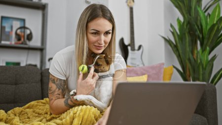 Téléchargez les photos : Une femme blonde joue avec son chien à l'intérieur, par un ordinateur portable, une guitare et des plantes. - en image libre de droit