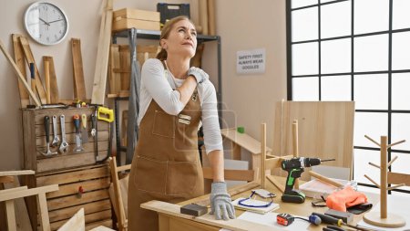 Photo for Mature woman in apron feeling shoulder pain while working in a carpentry workshop with tools around. - Royalty Free Image