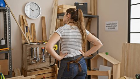 Photo for Back view of a young woman standing confidently in a well-equipped carpentry workshop. - Royalty Free Image