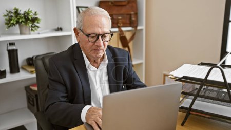 Foto de Hombre mayor inmerso en el éxito, trabajando intrincadamente con el ordenador portátil en su oficina de negocios, anciano profesional que muestra aguda concentración - Imagen libre de derechos