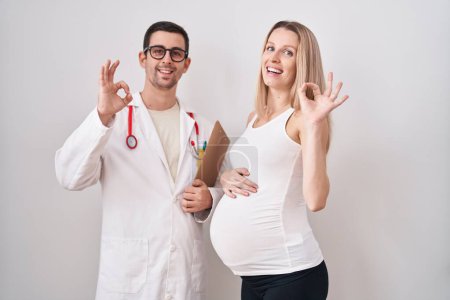 Foto de Mujer joven esperando un bebé con médico haciendo signo ok con los dedos, sonriendo gesto amistoso excelente símbolo - Imagen libre de derechos