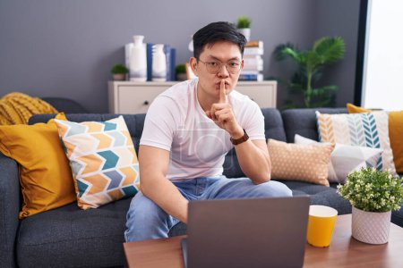 Téléchargez les photos : Jeune homme asiatique utilisant un ordinateur portable à la maison assis sur le canapé demandant à être calme avec le doigt sur les lèvres. silence et concept secret. - en image libre de droit