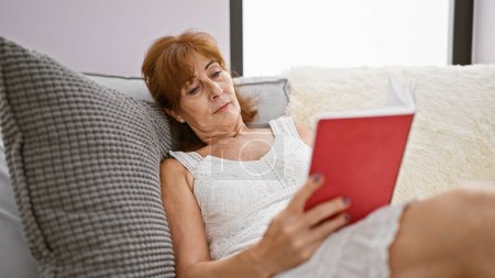 A mature woman relaxes at home on a couch, reading a red book in a cozy living room.