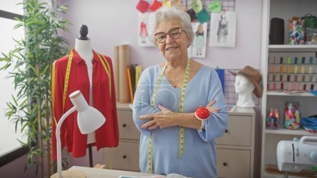 Photo for A smiling senior woman with arms crossed stands in a tailor shop surrounded by sewing materials, exuding confidence and expertise. - Royalty Free Image