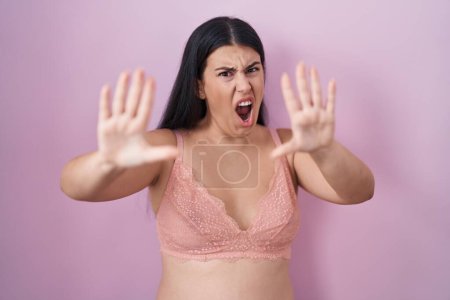 Foto de Mujer hispana joven con sujetador rosa asustada y aterrorizada con la expresión de miedo detener el gesto con las manos, gritando en shock. concepto de pánico. - Imagen libre de derechos