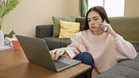 A young woman multitasking with a phone call and laptop in a cozy living room setting, appearing focused and casual.