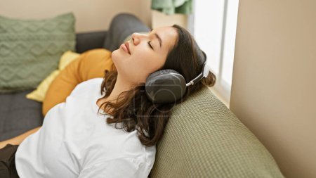 Photo for Young hispanic woman relaxing with headphones on a couch in a cozy living room - Royalty Free Image