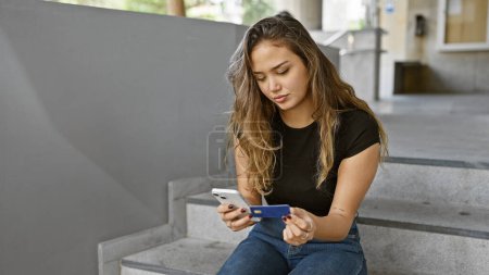 Photo for Young, beautiful hispanic woman hooking urban street lifestyle, seriously engaged in online shopping via smartphone, relaxed yet concentrated over secure credit card payment technology. - Royalty Free Image