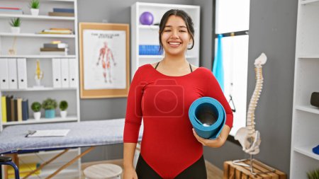Photo for A cheerful young hispanic woman holding a yoga mat poses in a well-equipped rehab clinic, indicating a healthy lifestyle and recovery. - Royalty Free Image
