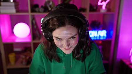 A caucasian woman in a gaming room with a headset smiles, lit by colorful neon lights at night.