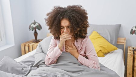 Young hispanic woman with curly hair looks unwell sitting on a bed, in a cozy bedroom setting at home.