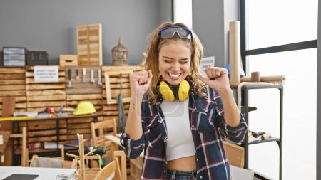 Photo for Vibrant celebration, young, beautiful hispanic woman carpenter revels in the win of her woodworking construction project, standing proudly in her workshop, safety glasses on - Royalty Free Image
