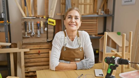 Téléchargez les photos : Femme caucasienne souriante portant un tablier dans un atelier de travail du bois avec des outils et un établi - en image libre de droit