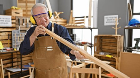 Ein widerstandsfähiger älterer Herr, ein erfahrener Schreiner, steht fest in seiner Tischlerei, setzt Sicherheitsbrillen auf und hält eine frisch gesägte Holzplanke in der Hand..