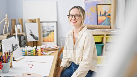 Foto de Joven mujer rubia artista sentado en la mesa sonriendo en el estudio de arte - Imagen libre de derechos