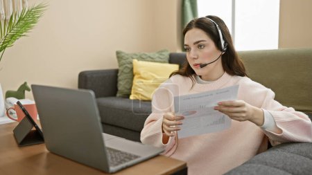 A focused woman wearing a headset reviews documents while working remotely on a laptop in a cozy living room.