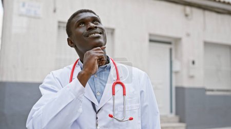 Un médico africano contemplativo con una bata blanca se encuentra fuera de un hospital, luciendo optimista.