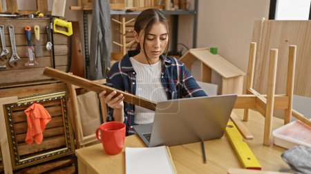 Téléchargez les photos : Une jeune femme hispanique examine un morceau de bois tout en travaillant sur un ordinateur portable dans un atelier de menuiserie rempli d'outils. - en image libre de droit