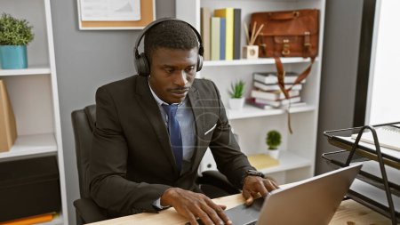 Foto de Empresario africano enfocado que usa auriculares mientras trabaja en una computadora portátil en un entorno de oficina moderno. - Imagen libre de derechos