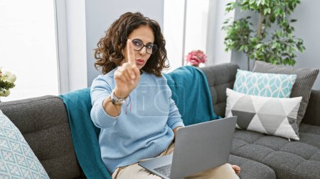 Hispanic woman with glasses gesturing indoors on a couch with a laptop, giving a stern look in a cozy home environment.