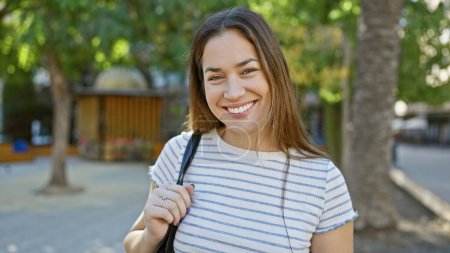 Téléchargez les photos : Jeune femme souriante aux cheveux longs bruns profitant de la lumière du soleil dans un parc verdoyant en plein air. - en image libre de droit