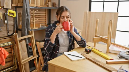 Foto de Una joven hispana disfruta de una pausa para tomar café en un taller de carpintería rodeado de herramientas y piezas de madera. - Imagen libre de derechos