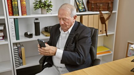Téléchargez les photos : Homme âgé prospère, sérieux mais détendu, conquérant monde des affaires, concentré sur le travail avec le téléphone, dactylographier des messages texte dans le bureau - en image libre de droit