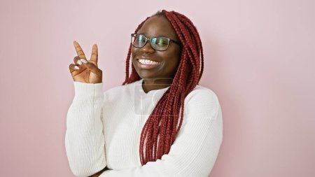 Téléchargez les photos : Portrait d'une femme afro-américaine souriante avec des tresses, faisant signe de paix sur un fond rose. - en image libre de droit