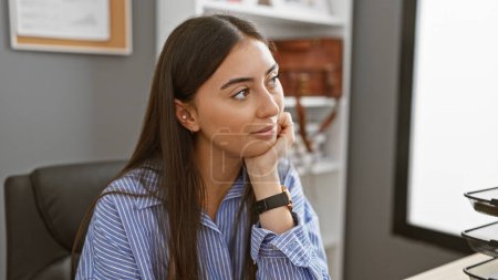Téléchargez les photos : Une jeune femme hispanique contemplative en chemise rayée pose dans un cadre de bureau moderne, respirant le professionnalisme et le calme. - en image libre de droit